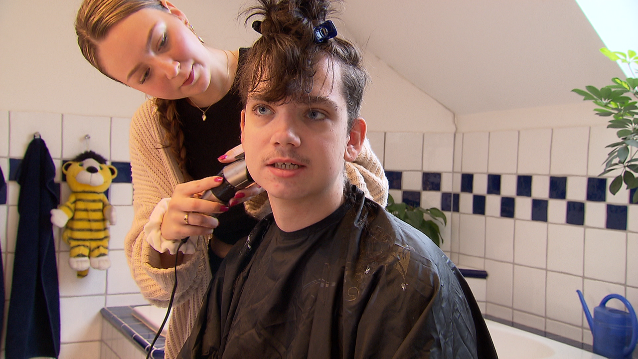 Marie schneidet mit einem Rasierer die Haare von Noah im Badezimmer.