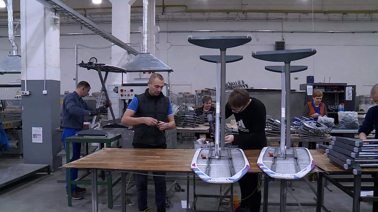Bügeltischfabrik von Innen. Recht im Bild zwei Bügelbretter auf dem Kopf gestellt, auf derm Tisch zur Bearbeitung in einer Halle.