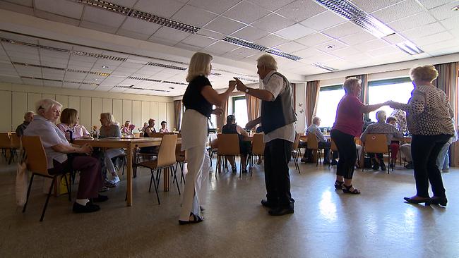 Pensionierte Menschen tanzen in einem kleinen Saal, daneben sitzen viele pensionierte Personen an Tischen.