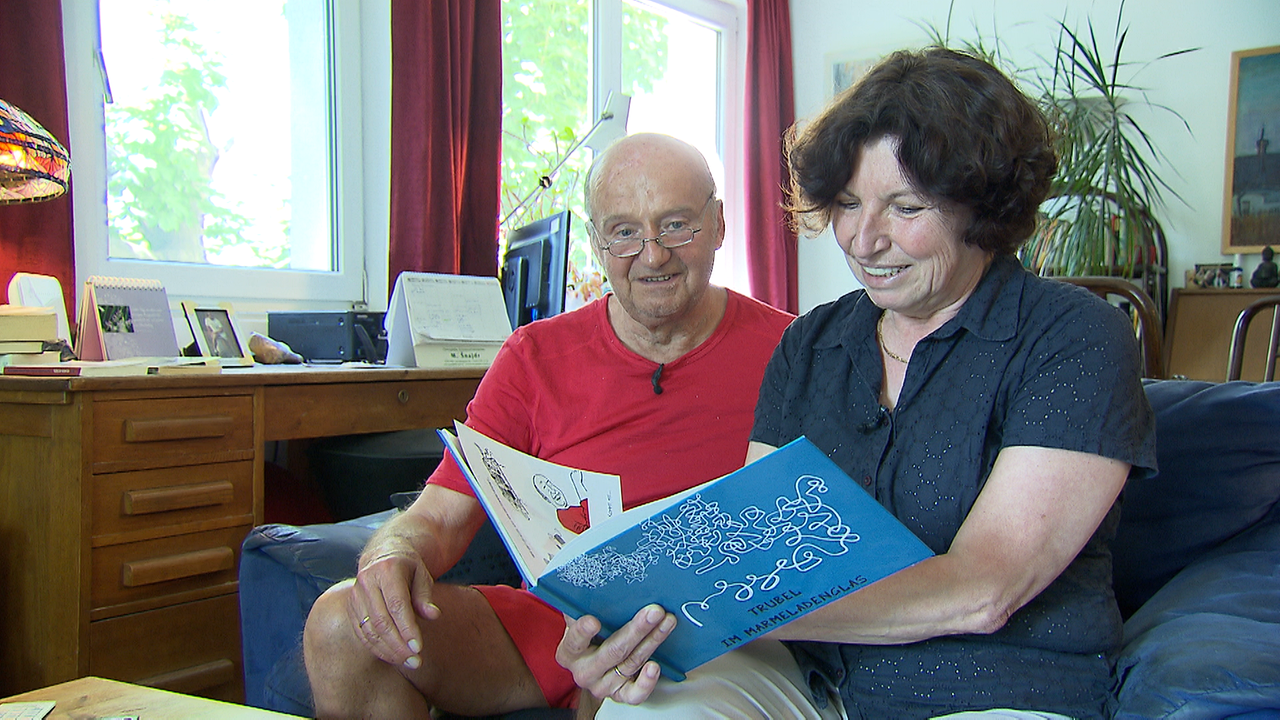 Andreas Trubel und seine Frau Beate im Wohnzimmer auf der Couch. Beate hat ein Buch in der Hand. 