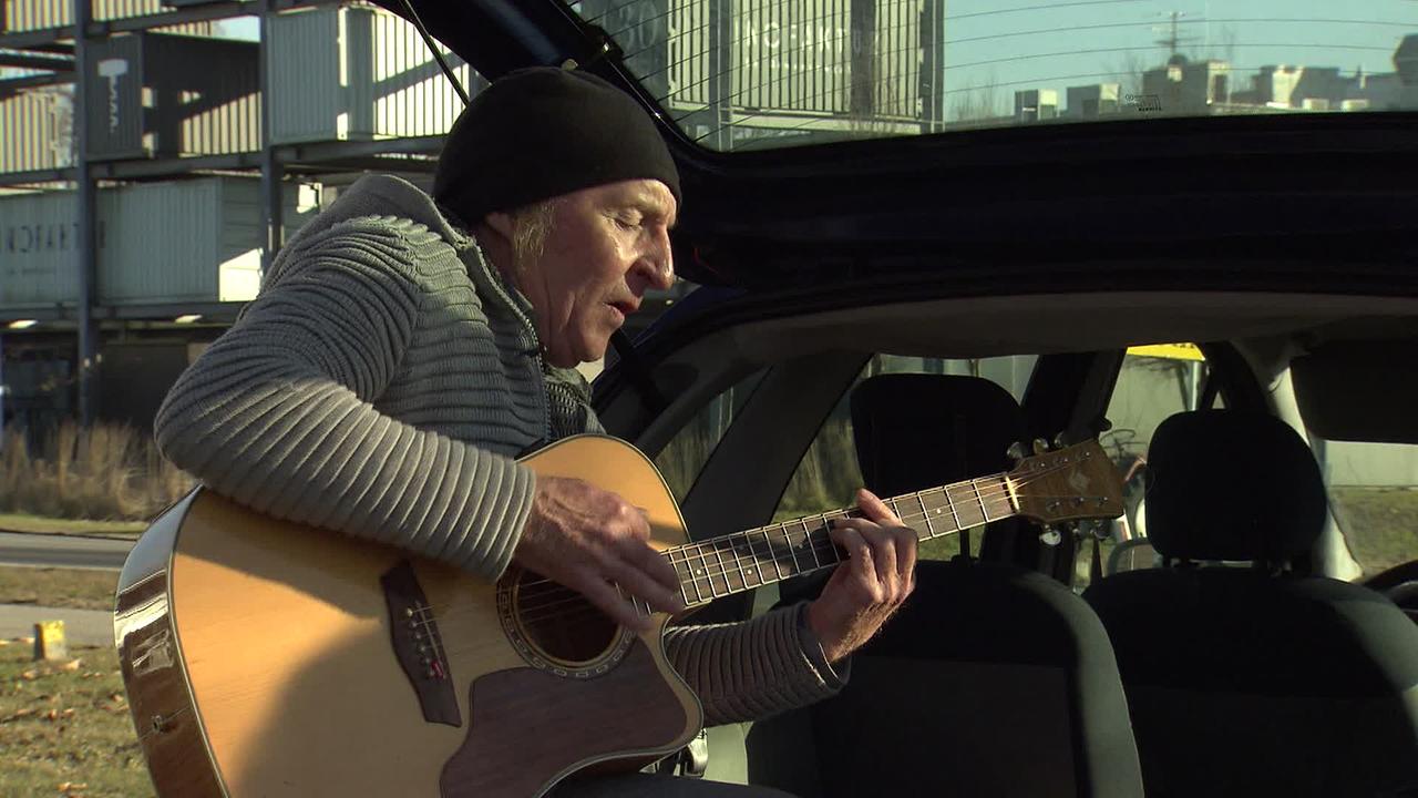 Herr M. steht beim offenen Kofferraum seines Autos und spielt auf seiner Gitarre und singt dazu.