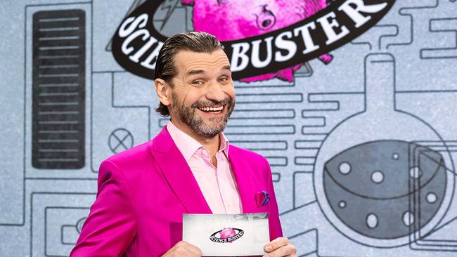 © ORF/Regine Schöttl Martin Puntigam im pinken Anzug steht vor dem „Science Busters“-Logo und hält eine Moderationskarte.