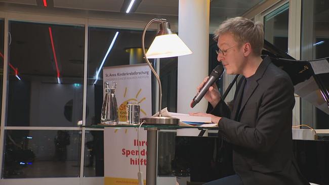 Schauspieler Max Müller sitzt an einem Pult vor dem Publikum und liest 