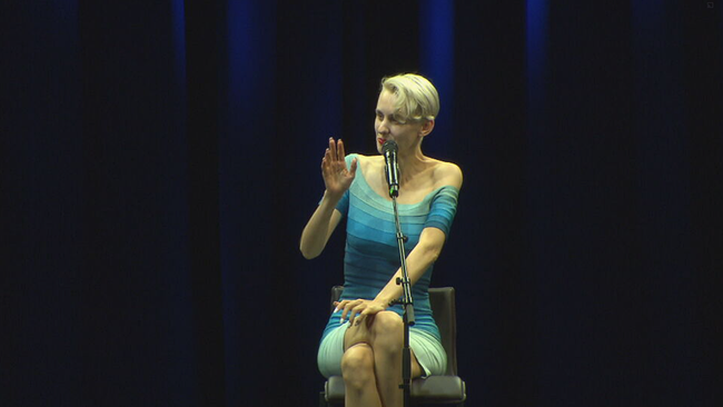 Lisa Eckhart sitzt in einem blau gestreiften Kleid auf der Bühne vor einem Mikrofon 