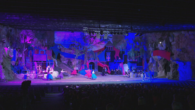 Blick auf die blau/rosa beleuchtete Kulisse des Steinbruch Winzendorf mit einigen Schauspielern in bunten Kostümen. 