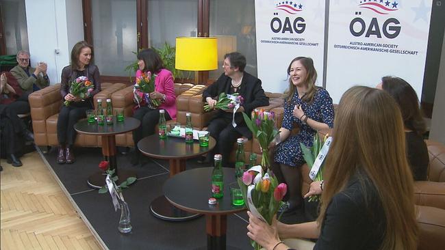 Sechs Frauen mit einem Tulpenstrauß in der Hand sitzen in Stühlen auf einem Podium