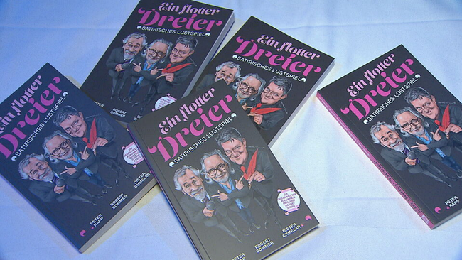 mehre Bücher mit dem Titel "Ein flotter Dreier" mit Karikatur-Figuren von Dieter Chmelar, Peter Rapp und Robert Sommer auf dem Cover liegen auf einem Tisch 