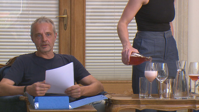 Thomas Maurer sitzt mit Unterlagen an einem Tisch vor Publikum. Nebem ihm wird gerade ein Glas Wein eingeschänkt.
