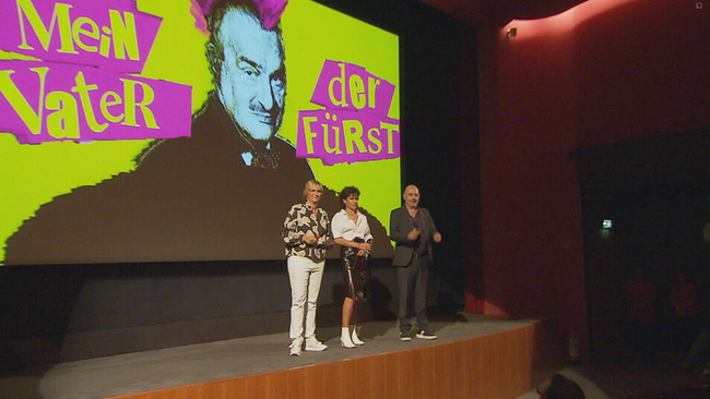 Lila Schwarzenberg steht mit zwei Moderatoren vor der Kinoleinwand auf der das Plakat zum Film zu sehen ist