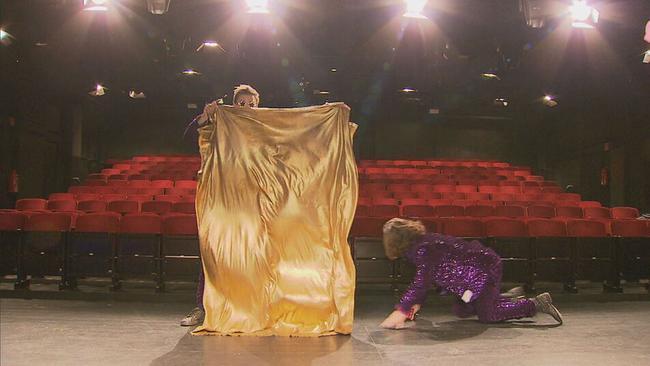 Siegfried & Joy auf der Bühne. Einer hält ein goldenes Tuch in der Hand, der andere krabbelt in einem Glitzeranzug auf dem Boden