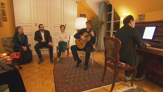Mijou Kovacs und ein Teil ihrer Gäste sitzen in einem Wohnzimmer und lauschen einem Klavier- und einem Gitarrespieler 