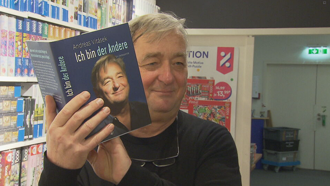 Andreas Vitásek hält sein Buch "Ich bin der Andere" in die Kamera und lächelt 