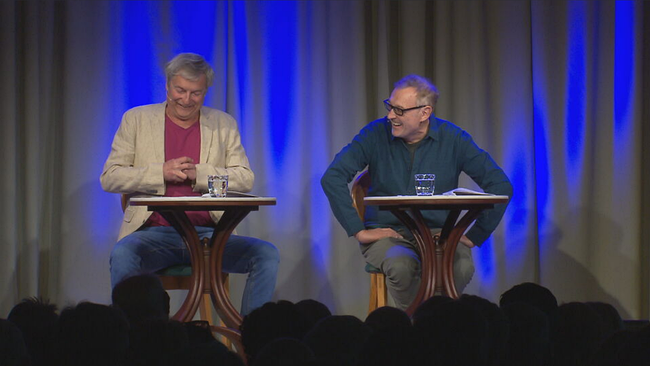Josef Hader und Alfred Dorfer  sitzen mit einem Tischchen vor sich auf der Bühne und lesen aus einem Mnuskript vor. Beide lachen.  