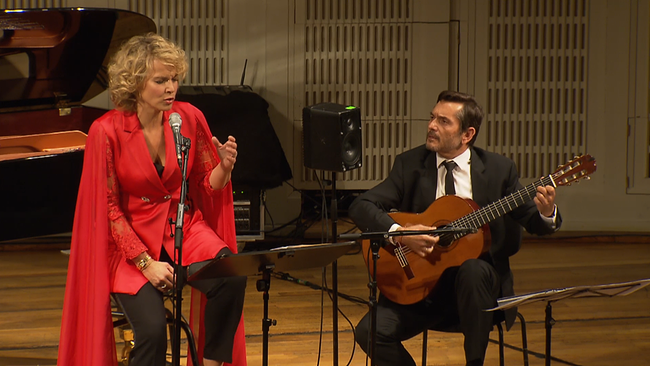 Opernsängerin Elīna Garanča sitzt in einem roten, langen Blazer neben José María Gallardo del Rey, der sie mit der Gitarre begleitet
