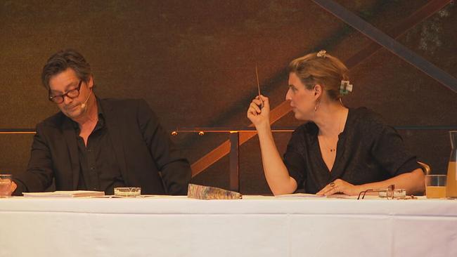Elena Uhlig und Fritz Karl sitzen an einem Tisch auf der Bühne. Sie zeigt mit einem Stift auf ihn