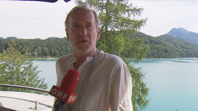 Schauspieler Peter Lohmeyer mit ORF Mikrofon in einer Interviewsituation. Im Hintergrund der Fuschlsee 