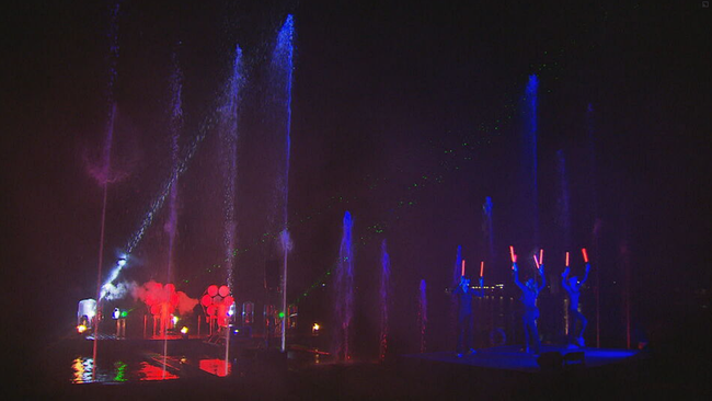 Künstler mit Leuchstäben auf einer Bühne mit Wasserfontänen und leuchtenden Luftballons 