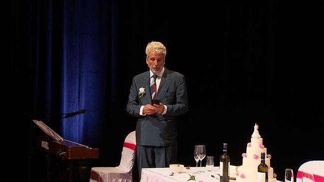Dirk Stermann steht im Anzug auf der Bühne. Vor ihm steht eine gedeckte Tafel mit Weinflaschen und einer Hochzeitstorte 