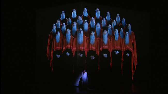 Ève-Maud Hubeaux als Amneris auf der Bühne. Hinter ihr: eine Gruppe von Männern mit langen, weißen Bärten, schwarzem Hut und rot-schwarzen Roben