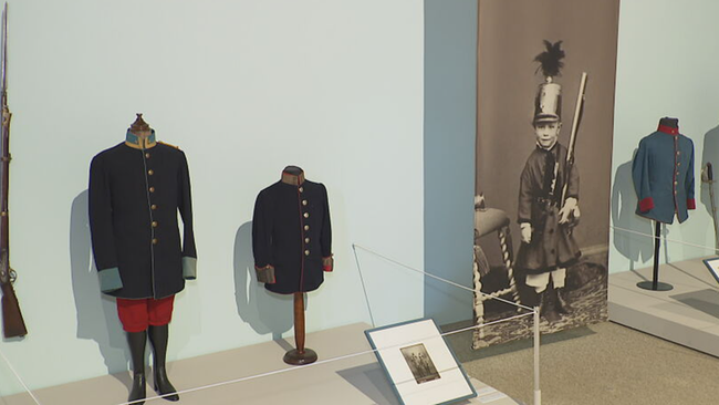 Blick in die Ausstellung: Uniformen, Gewehre neben Schwarzweiß-Fotos von Kindern die damit posieren