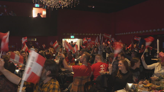 Publikumsraum im Vindobona. Die viele Gäste sitzen an Tischen und winken mit österreichischen Flaggen.