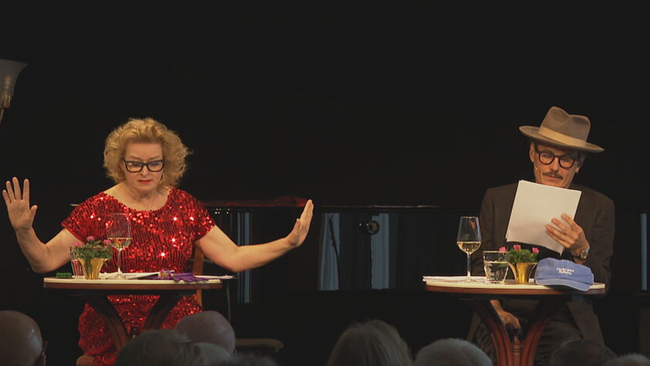 © ORF / Seitenblicke Petra Morzé und Manuel Rubey sitzend auf einer Bühne, mit Tischen, Texten, Wein und Blümchen