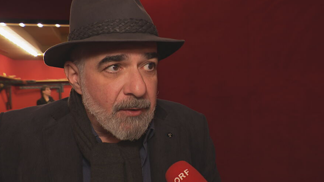 Michael Niavarani (in schwarzer Kleidung und mit schwarzem Hut) in der Interview-Situation mit Mikro vor dem Mund