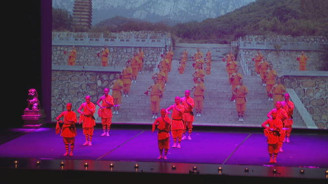 9 Mönche in drei Dreier-Reihen stehen auf der Bühne in traditionellem Gewand
