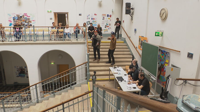 Aula mit Besucherinnen und Besuchern, Vortragende in der Mitte der Stufen an einem Tisch