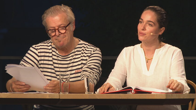 Koch (in schwarz weiß gestreiftem Shirt) und Altenberger (in weißer Bluse) sitzen am Tisch und lesen, dahinter dunkle Nacht. 