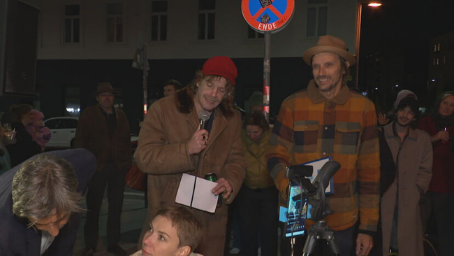 Voodoo Jürgens und Skero stehen mit Mikrofon in der Hand nachts auf Gehsteig in Gruppe von Leuten