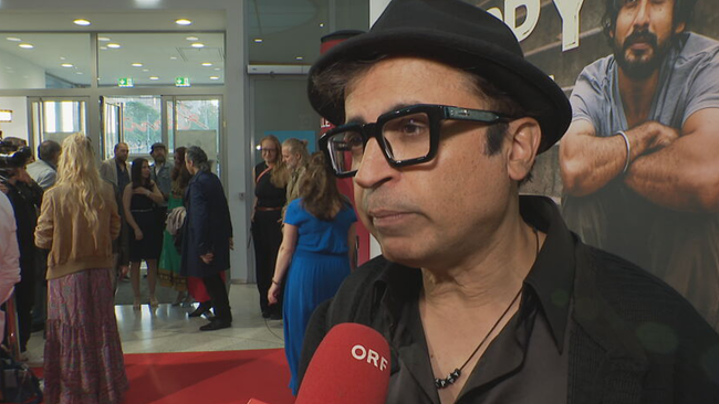 © ORF / Seitenblicke Regisseur steht vor Film-Plakat und gibt Interview. Im Hintergrund geben andere Prominente Interviews und werden fotografiert.