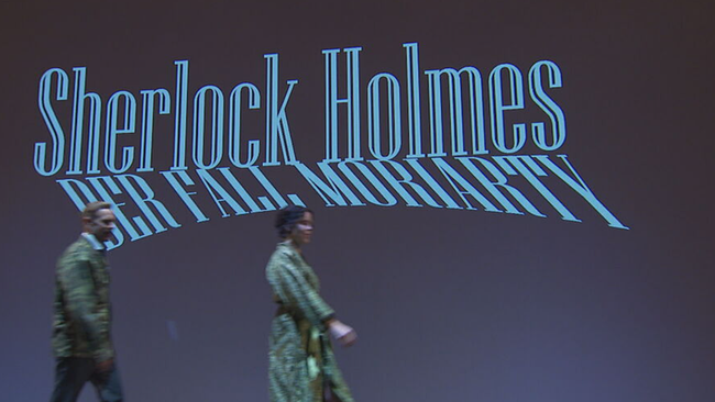 An der Wand steht der Schriftzug "Sherlock Holmes - Der Fall Moriarty" und davor gehen eine Darstellerin und ein Darsteller auf der Bühne von links nach rechts