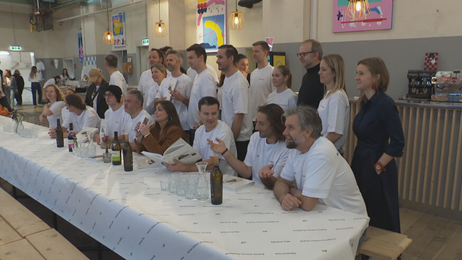 Gastronomen (manche mit Kochbuch) sitzen an und stehen hinter Tisch, und schauen teilweise zum Fotografen.