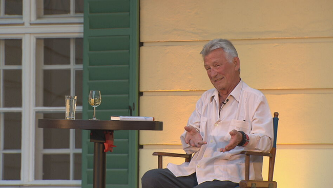 Heinz Marecek gestikuliert auf der Bühne sitzend mit seinem Buch und einem Glas Wein neben sich 