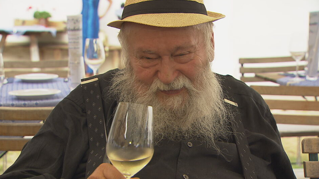 Herman Nitsch prostet lächelnd mit einem Glas Weißwein Richtung Kamera