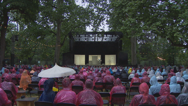 Publikum im Belvedere-Park in rosa und blauen Regenponchos schauen auf die überdachte Bühne