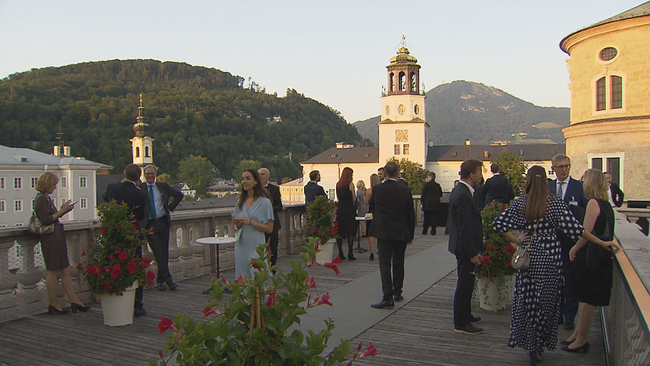 Gäste auf einer Dachterrasse in Gesprächen mit Blick auf die innere Stadt Salzburgs im Abendlicht