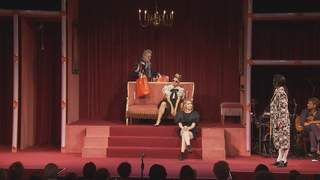 rotsamtenes Bühnenbild, die SchauspielerInnen sitzen und stehen versetzt auf Couch und Treppe