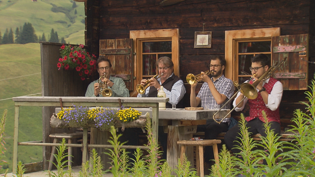 Vier Männer vor einer Berghütte sitzend spielen Blasinstrumente