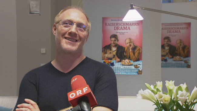 Simon Schwarz beim Interview mit verschränkten Armen, lächelnd vor dem Filmposter