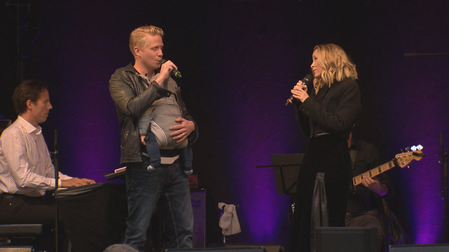 Missy May mit Ehemann Andreas Wanasek, der ein schlafendes Baby umgeschnallt hat, singend auf der Bühne