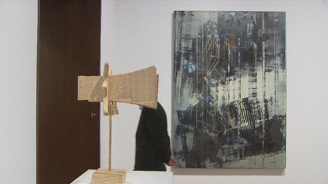 Foto der Kunstausstellung: Rechts ein hell-dunkelgraues Ölgemälde, ein Mann läuft daran vorbei und ist hinter einer Skulptur aus Zeitungspapier versteckt