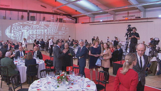 Raum voller Menschen in Business-Kleidung, Kameras, gedeckte runde Tische, Apa-Logo-Projektion im Hintergrund