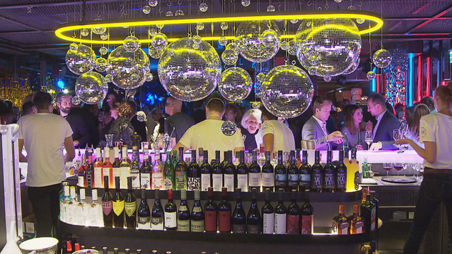 Bar mit großer Auswahl an (Wein-)Flaschen, darüber viele Discokugeln in verschiedenen Größen, hinter der Bar, Clubgäste