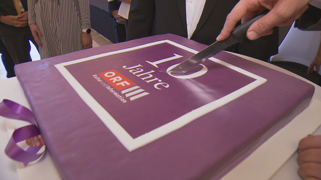 Großer viereckiger, lila Geburtstagskuchen wird angeschnitten, es ist zu lesen "10 Jahre ORF3"