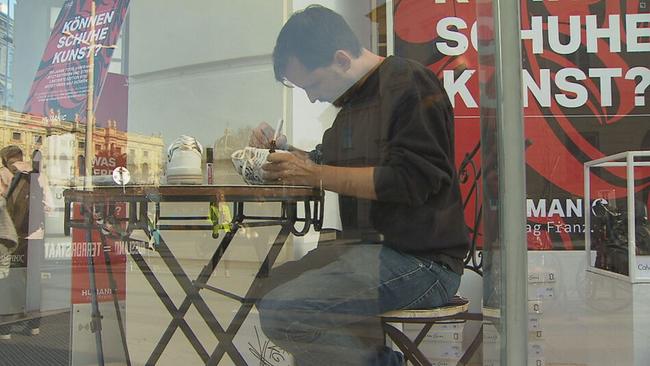 Streetart-Künstler Fresh Max sitzt in einem Schaufenster und bemahlt weiße Sneaker mit einem schwarzen Edding 