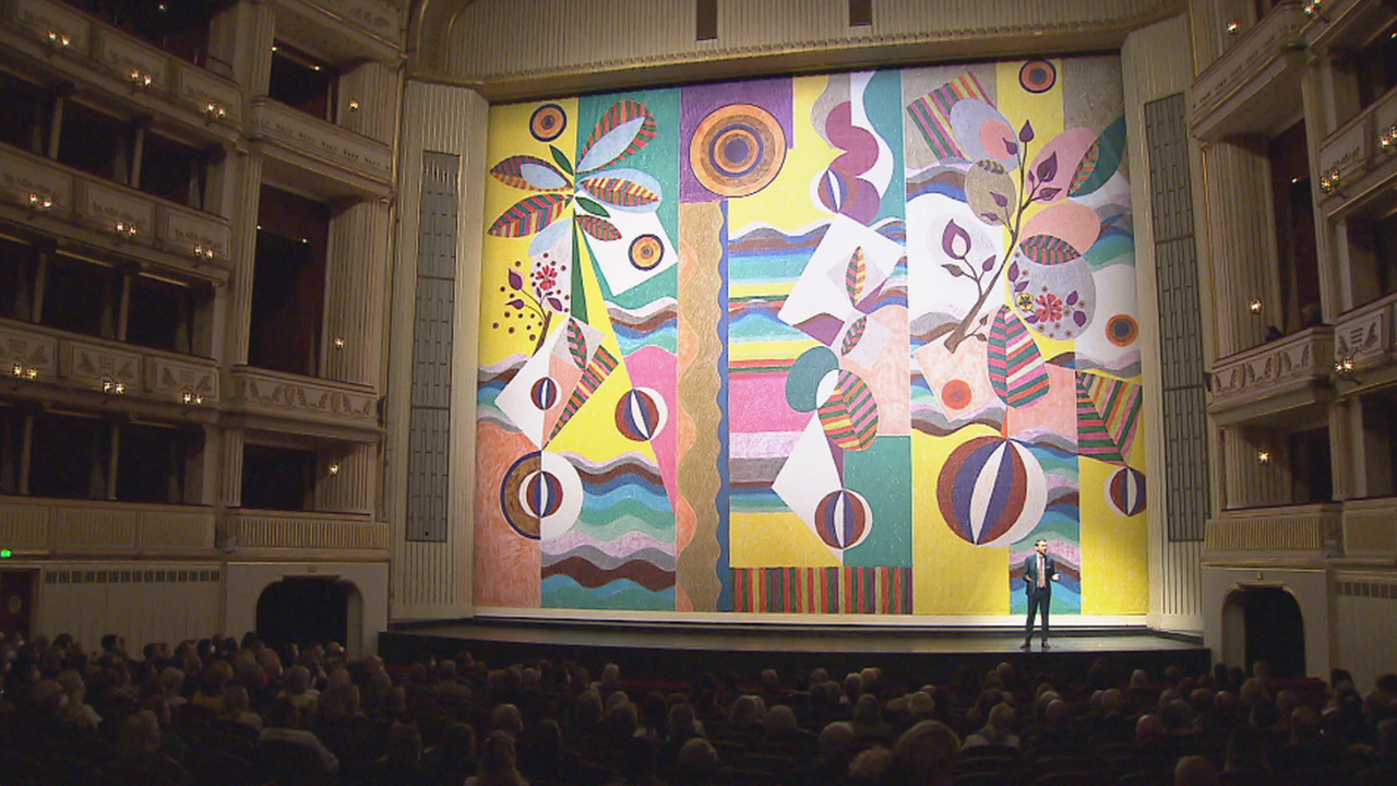 Blick aus dem Publikum auf die Bühne, links und rechts Ränge, der eiserne Vorhang ist bunt gemustert, viele Kontraste in Farben und Formen