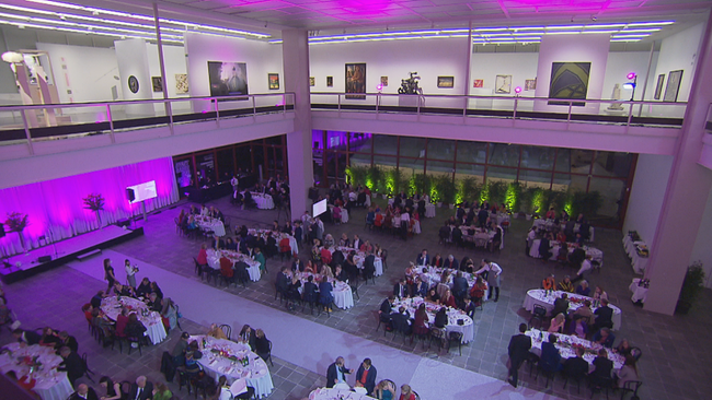 Totale über das offene Erdgeschoss mit ovalen, voll besetzten Tischen, im 1. Stock die leeren Ausstellungsräume und Bilder, alles in lila Licht getaucht