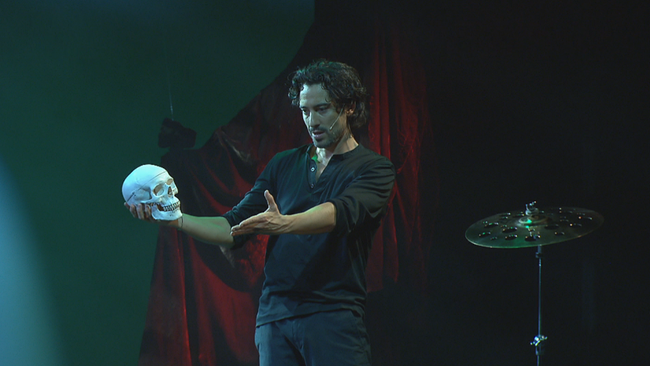 Bernardin auf der Bühne mit einem Totenschädel in der einen Hand, die andere ausgestreckt, dunkler Hintergrund, düstere Stimmung